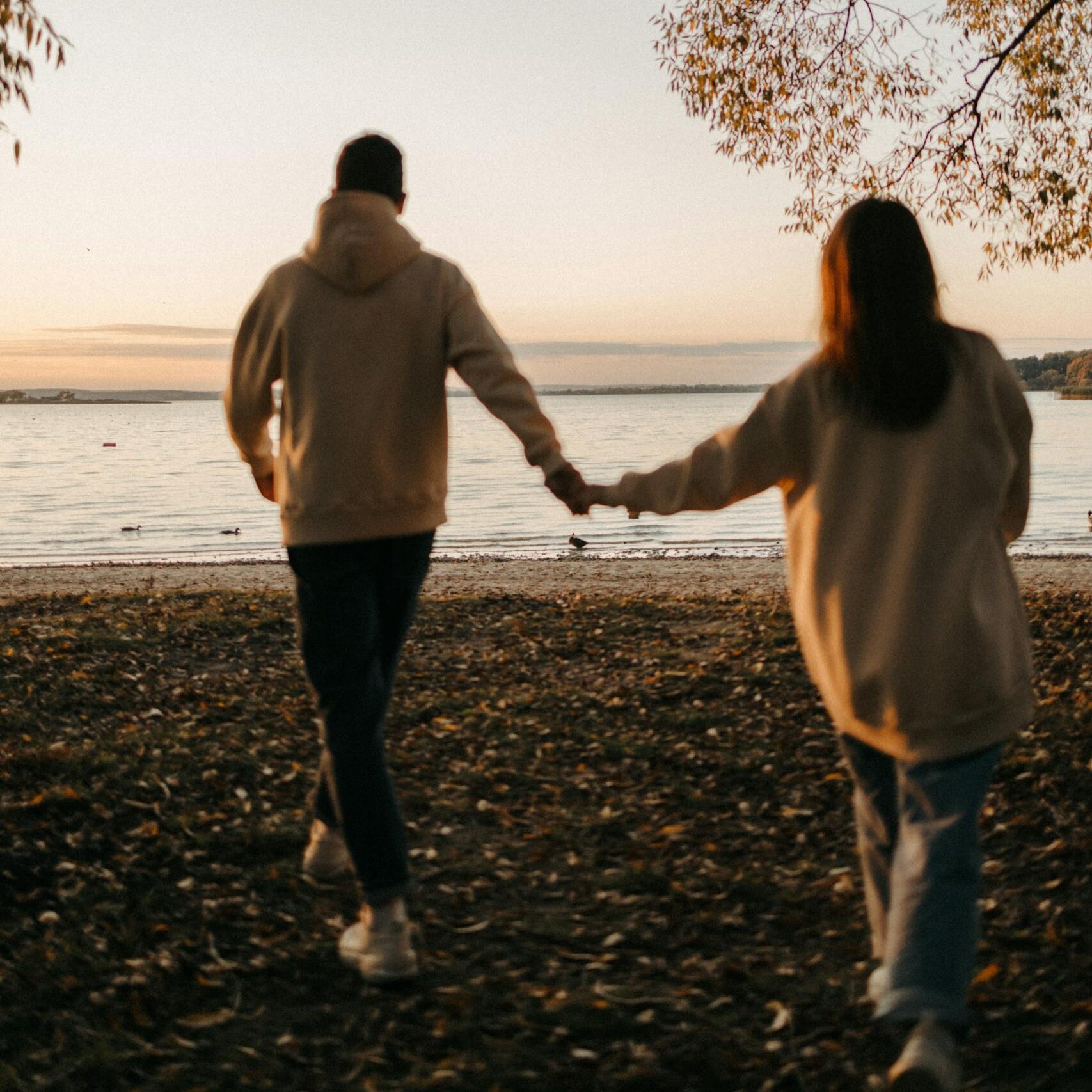 A romantic couple holding hands while walking towards the sunset by the beach, surrounded by autumn scenery.